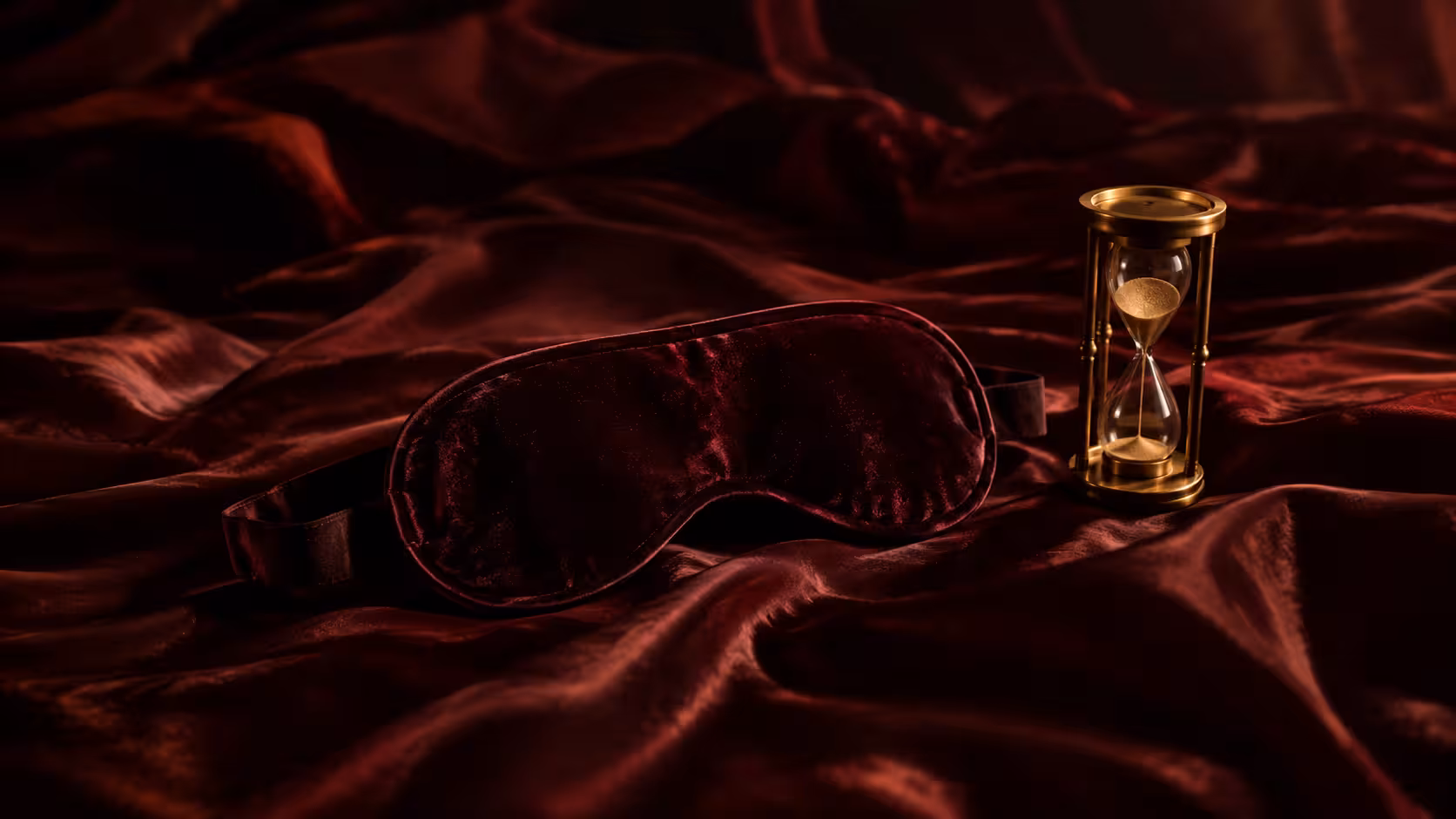 Venda de veludo sobre lençóis de cetim bordô ao lado de uma ampulheta, simbolizando o controle de orgasmo, a expectativa e o adiamento do prazer no BDSM.
